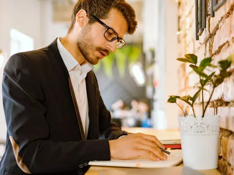 A man in glasses writing notes at a bright cafe table beside a potted plant