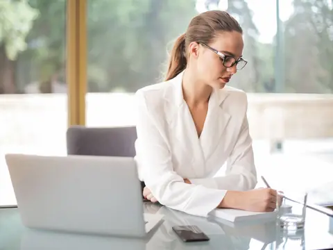 A woman writing notes beside her laptop at a desk by a bright window