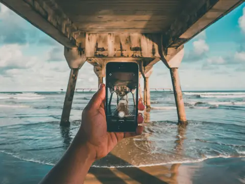 A hand holding up a phone under a wooden pier at the water's edge on a bright day