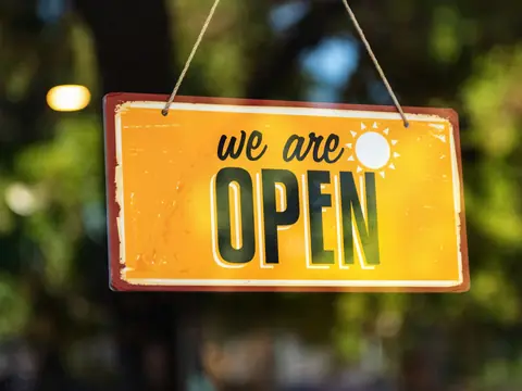 A yellow We Are Open sign hanging outside a shop in the sun