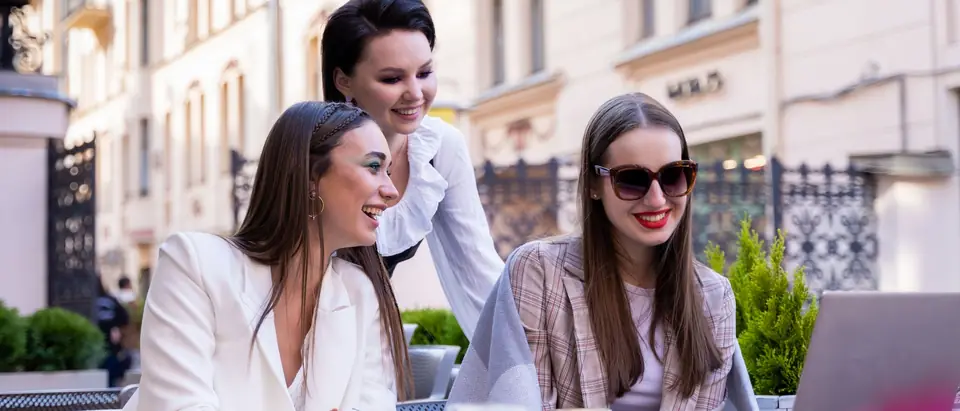Three women working on a laptop at a sunny outdoor cafe table