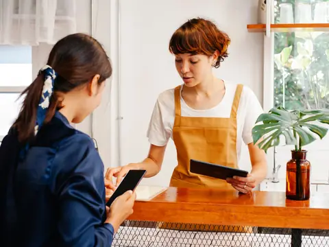 A customer holding her phone at the counter of a bright cafe while the owner checks a tablet