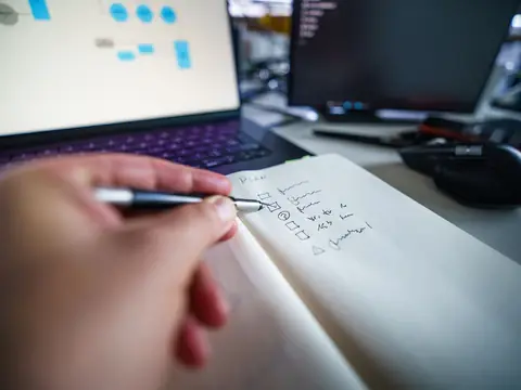 A hand ticking off a written checklist beside a laptop