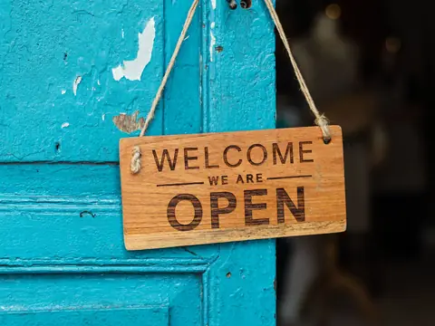 A wooden Welcome, We Are Open sign hanging on a turquoise shop door
