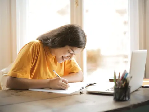 A woman smiling as she writes notes at a sunny desk by a window