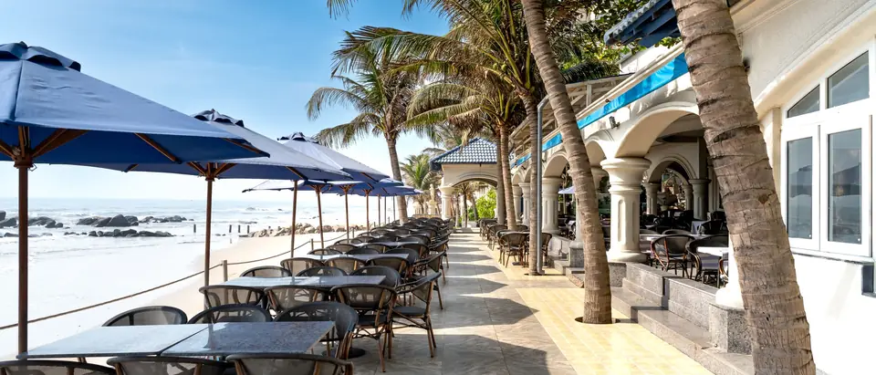 A beachfront restaurant patio with blue umbrellas and palm trees on a sunny day