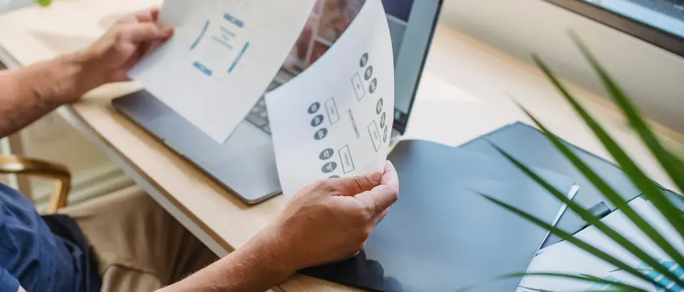 A person holding printed website wireframes beside a laptop at a bright desk